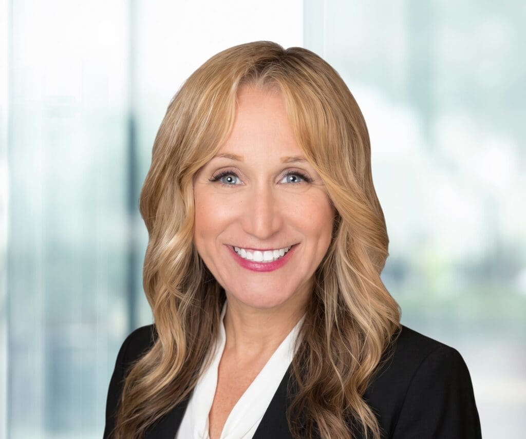A woman with long blonde wavy hair, wearing a black blazer and white blouse, smiles at the camera. The background is softly blurred with natural light coming through large windows.