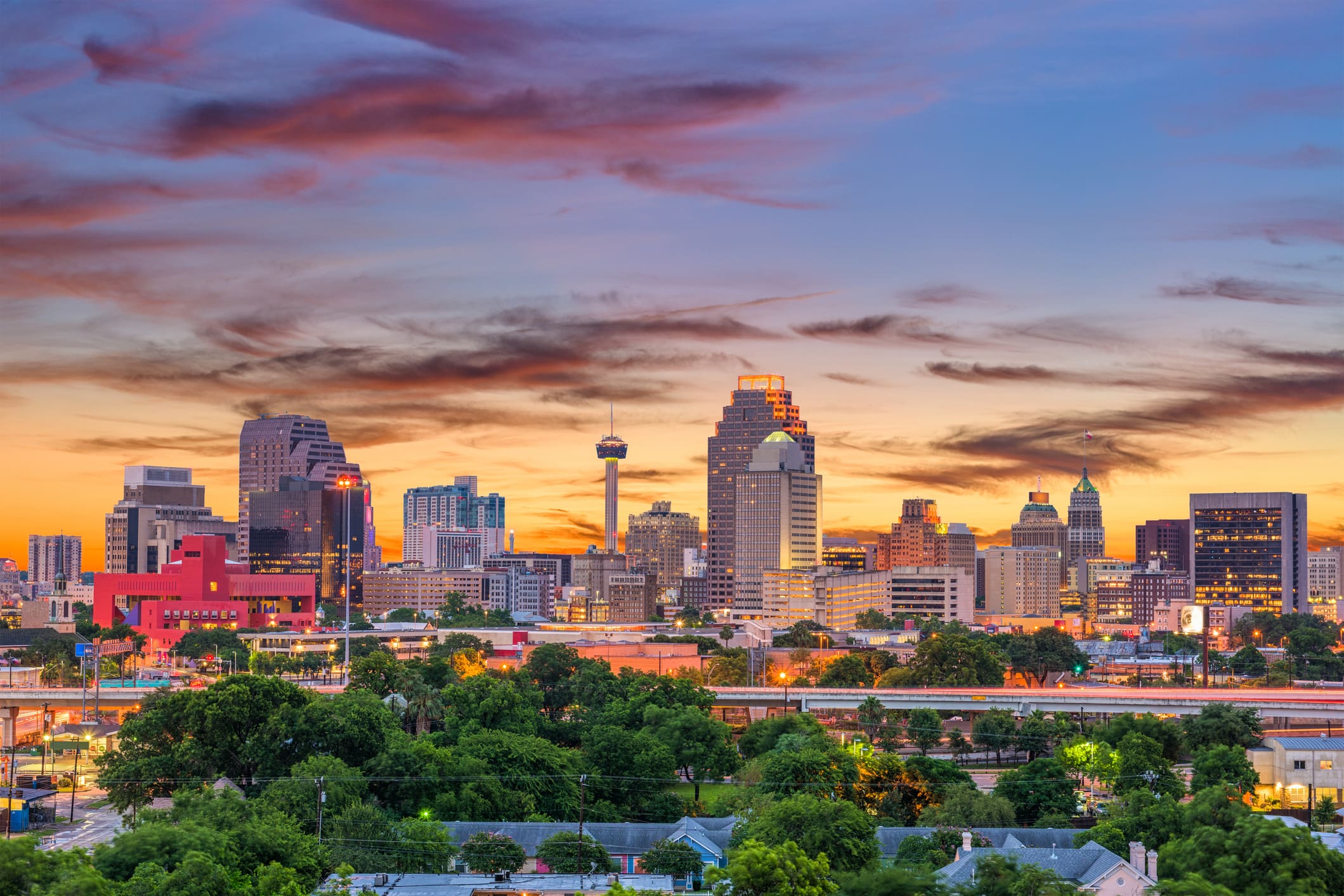 San Antonio city skyline at sunset with colorful clouds, modern downtown buildings, and lush green trees in the foreground.